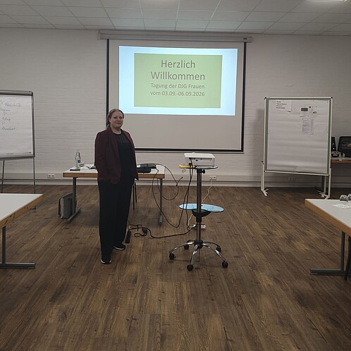 Eine Frau steht in einem Smeinarraum neben einem Beamer. Im Hintergrund läuft eine Powerpoint-Präsenation