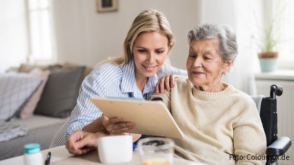 Eine junge Frau sitzt mit einer älteren Frau im Rollstuhl am Esstisch und hilft ihr am Tablet.