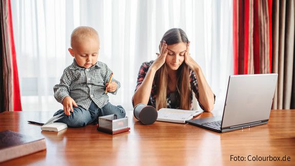 Eine sichtlich gestresste Mutter versucht zu Hause am Laptop zu arbeiten, während neben ihr auf dem Schreibtisch ein Kleinkind sitzt und mit Büroutensilien spielt.
