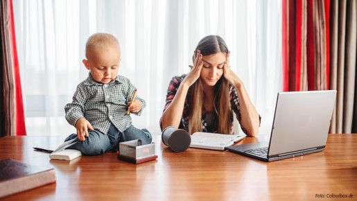 Eine sichtlich gestresste Mutter versucht zu Hause am Laptop zu arbeiten, während neben ihr auf dem Schreibtisch ein Kleinkind sitzt und mit Büroutensilien spielt.