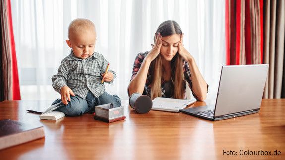 Eine sichtlich gestresste Mutter versucht zu Hause am Laptop zu arbeiten, während neben ihr auf dem Schreibtisch ein Kleinkind sitzt und mit Büroutensilien spielt.