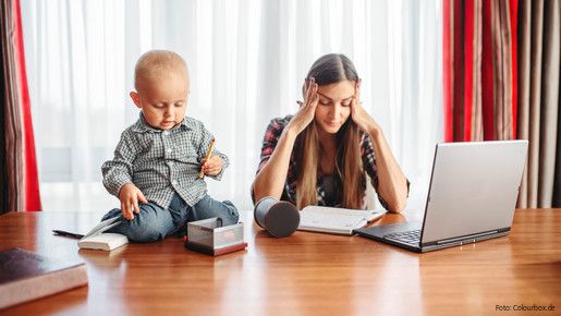 Eine sichtlich gestresste Mutter versucht zu Hause am Laptop zu arbeiten, während neben ihr auf dem Schreibtisch ein Kleinkind sitzt und mit Büroutensilien spielt.