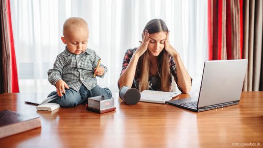 Eine sichtlich gestresste Mutter versucht zu Hause am Laptop zu arbeiten, während neben ihr auf dem Schreibtisch ein Kleinkind sitzt und mit Büroutensilien spielt.