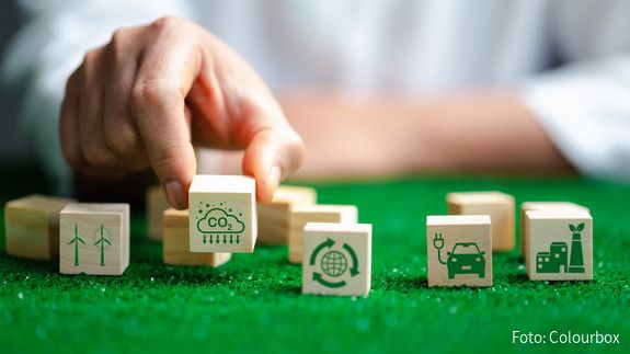 Nahaufnahme einer Hand, die auf einer grünen Fläche verschiedene Würfel mit Symbolen für erneuerbare Energien platziert. 