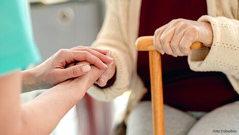 Das Foto zeigt die Hände einer Pflegerin, die die Hände einer Seniorin mit einem Stock halten. Es handelt sich um eine Nahaufnahme.
