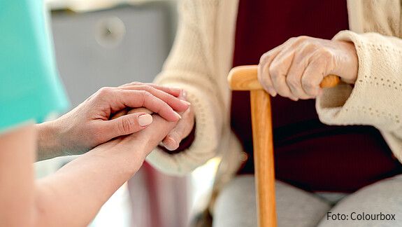 Das Foto zeigt die Hände einer Pflegerin, die die Hände einer Seniorin mit einem Stock halten. Es handelt sich um eine Nahaufnahme.