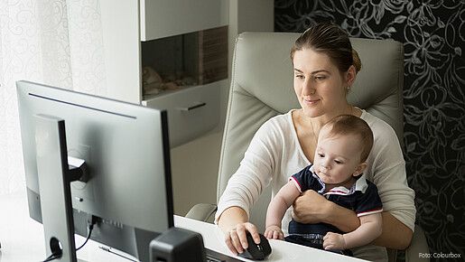 Zu sehen ist eine junge Mutter, die am Schreibtisch Homeoffice macht und ihren kleinen Sohn auf dem Arm hält.