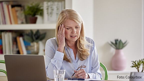 Eine Frau in den Wechseljahren sitzt an einem Schreibtisch und hat Kopfschmerzen