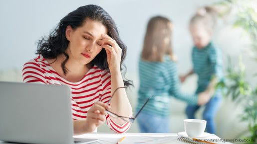 Eine dunkelhaarige Frau arbeitet am Laptop und reibt sich gestresst die Augen, im Hintergrund spielen zwei kleine Mädchen