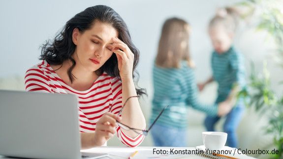 Sorgearbeit fair teilen Eine dunkelhaarige Frau arbeitet am Laptop und reibt sich gestresst die Augen, im Hintergrund spielen zwei kleine Mädchen