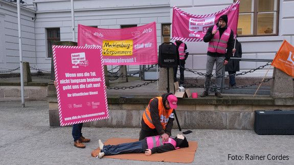 Fahrrad Demo Schwerin Eine Gruppe von Menschen ist am protestieren. Im Mittelpunkt übt jemand eine Herzdruckmassage an einer Puppe aus. Daneben ist ein Plakat zu sehen, auf dem steht: Wenn uns die TdL nicht langsam beatmet geht uns die Luft aus!