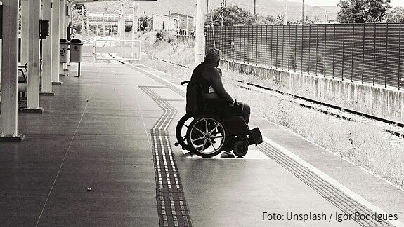 Schwarz-weiß-Foto eines Mannes, der an einem Bahnsteig in einem Rollstuhl sitzt und auf den Zug wartet.