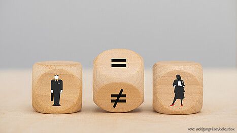 Zu sehen sind drei Würfel: Einer mit einer männlichen Figur, einer mit einer weiblichen. Der Würfel in der Mitte steht gekippt, ein Gleichheitszeichen und ein Ungleichheitszeichen sind zu sehen.
