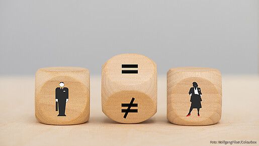 Zu sehen sind drei Würfel: Einer mit einer männlichen Figur, einer mit einer weiblichen. Der Würfel in der Mitte steht gekippt, ein Gleichheitszeichen und ein Ungleichheitszeichen sind zu sehen.