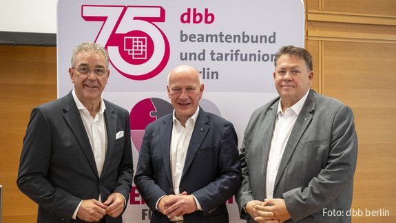 Drei Männer in Anzügen stehen vor einem Aufsteller und blicken in die Kamera