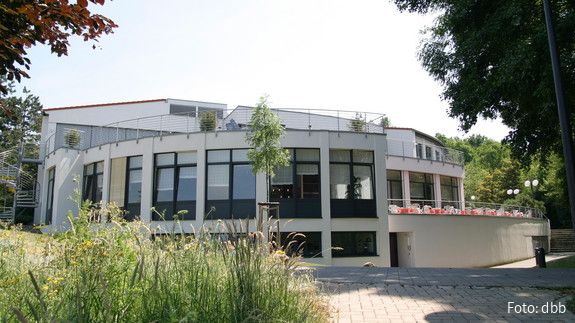 Pflegeseminar der dbb bundesseniorenvertretung Ein großes, helles, rundliches Haus ist im Mittelpunkt des Bildes. Im linken Vordergrund ist eine hochwachsende Wiese zu sehen.