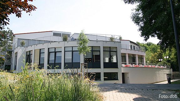 Ein großes, helles, rundliches Haus ist im Mittelpunkt des Bildes. Im linken Vordergrund ist eine hochwachsende Wiese zu sehen.