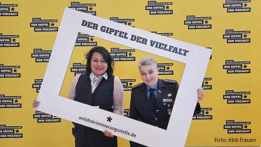 Zwei Frauen halten einen großen weißen Bilderrahmen vor einer gelben Wand