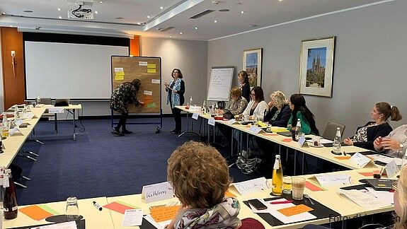 Blick von hinten in einen Konferenzraum. Frauen sitzen an Tischen in U-Form und blicken nach vorne.