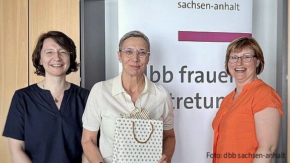 Zwei Frauen überreichen einer dritten Frau eine Geschenktüte