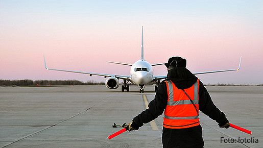 Ein Fluglotse in einer roten Warnweste steht mit dem Rücken zur Kamera. Weit vor ihm ist ein Flugzeug zu sehen, das auf der Flugbahn steht.